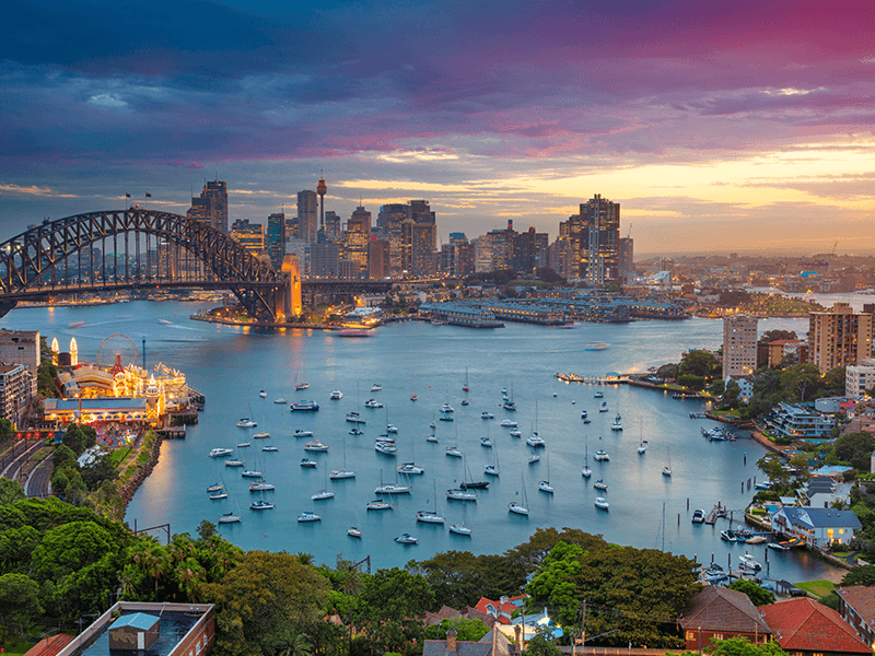view of sydney harbour bridge, luna park and sydney harbor with buildings and sailboats at sunset