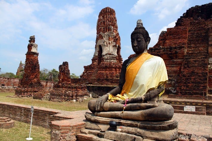 sitting buddha draped in orange sash in the middle of ruins