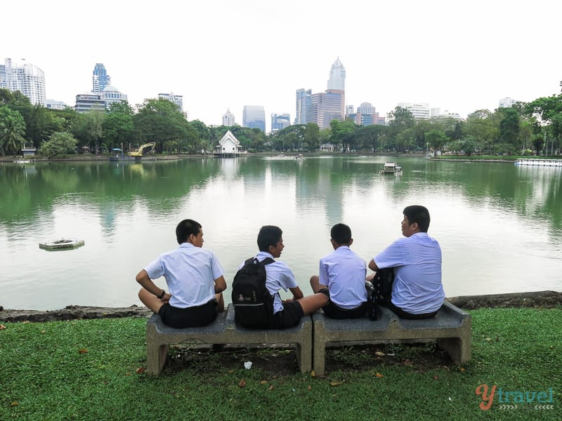 school boys sitting on bench looking at view of lake and buildings at lumphini park