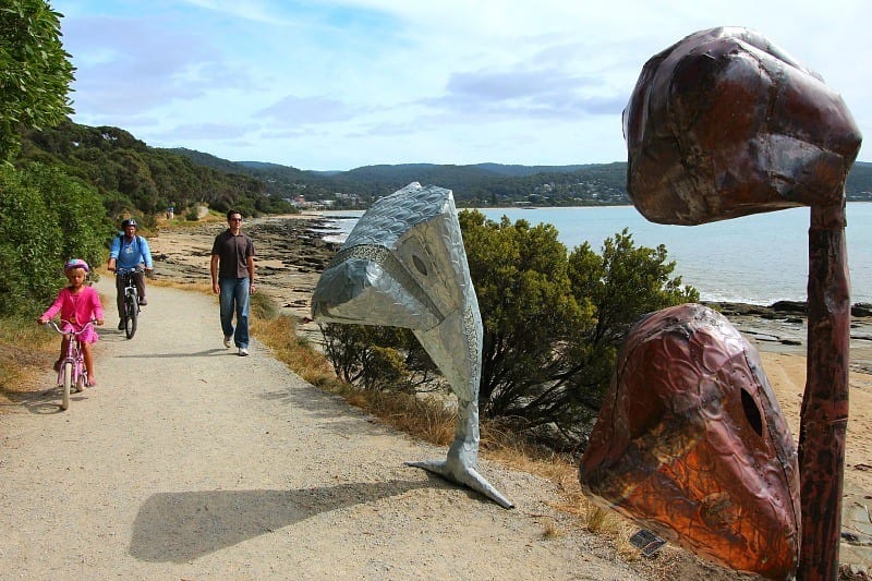 people riding past sculptures on dirt trail beside ocean