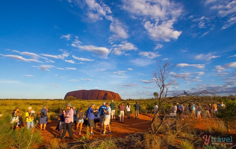 people standing on a dirt path looking at a large red rock