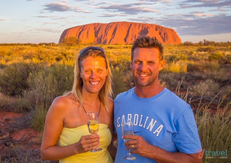 people smiling and holding drinks in front of uluru at sunset