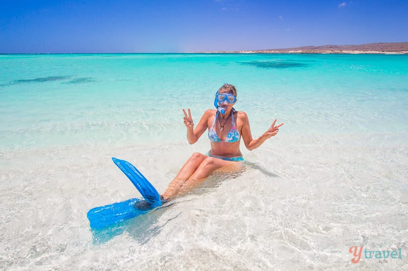 woman with snorkels on, posing with peace signs to the camera while sitting in water. 