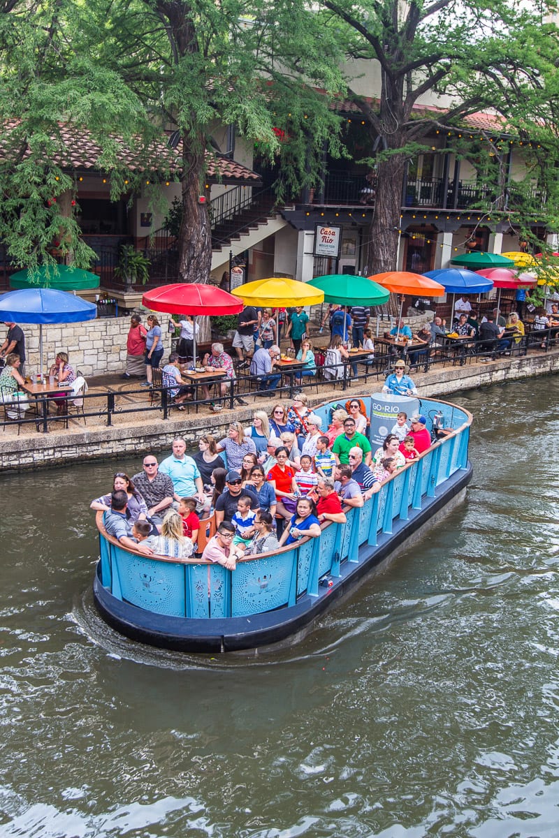 tables with colorful umbrellas on riverwalk and boats going by on river