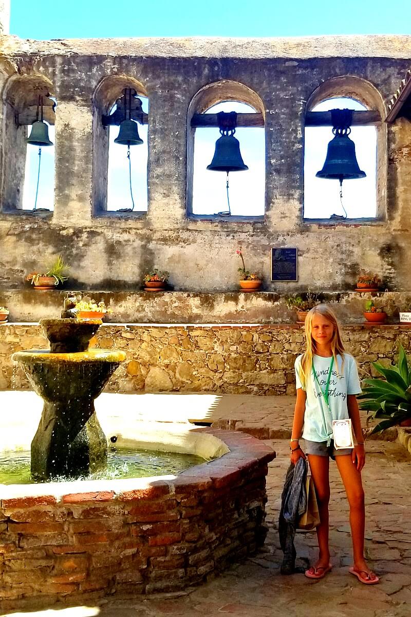 girl standing beside fountain with bells in the background at Mission San Juan Capistrano