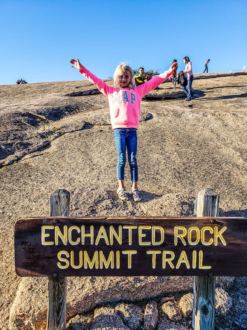 girl with arms in the air behind trail sign that says Enchanted Roc summit trail