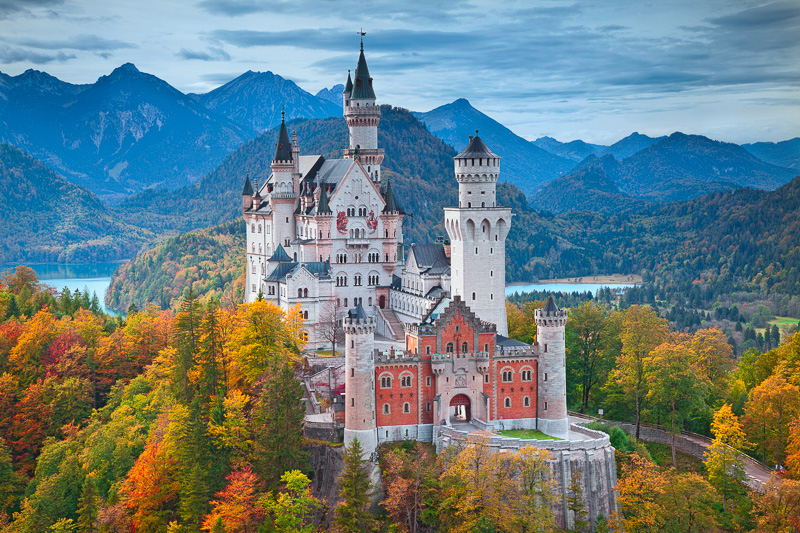 Stunning fairytale Neuschwanstein castle on a small hill surrounded by Fall folage with a lake and mountain ranges in the background