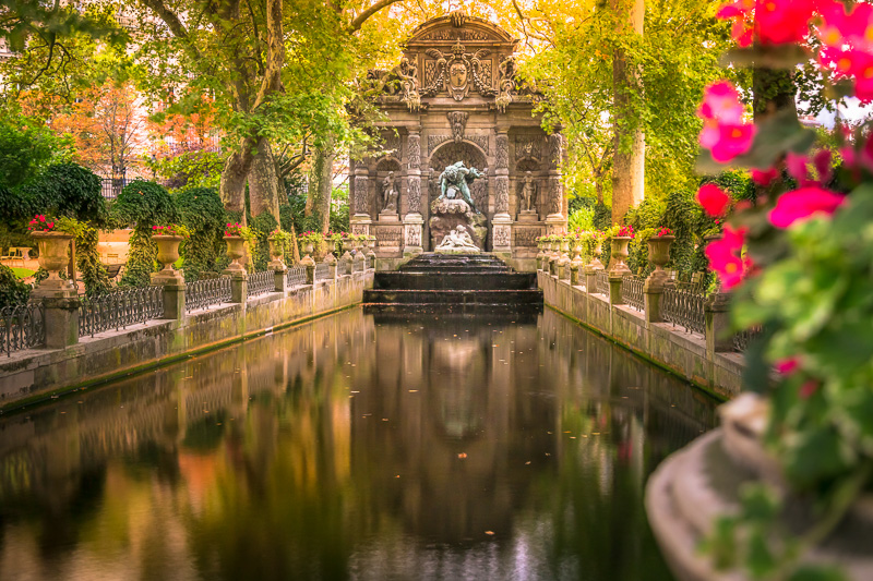 Fountain pond surrounded by lush greenish  trees and agleam  pinkish  flowers