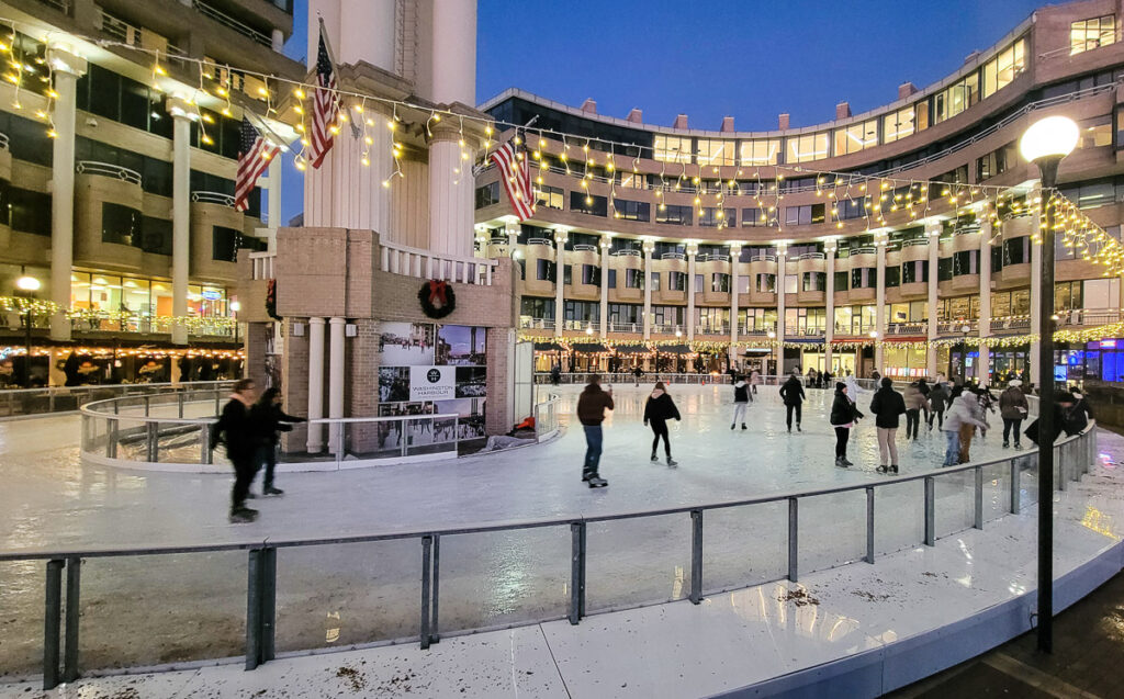People ice skating inside a rink surrounded by buildings
