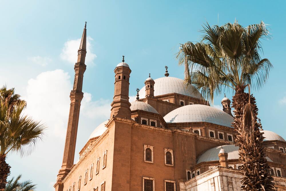 brown mosque with white domed roofs and palm trees framing it