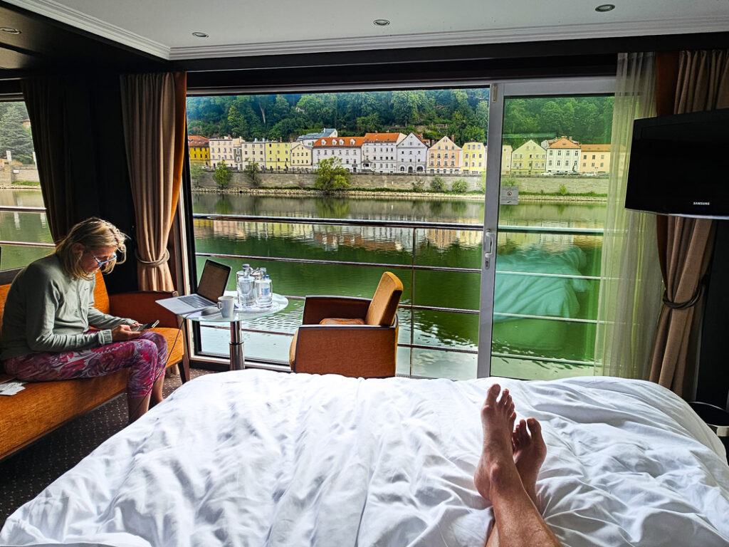 Legs on a bed and lady sitting on couch with views of a river from their room on a cruise