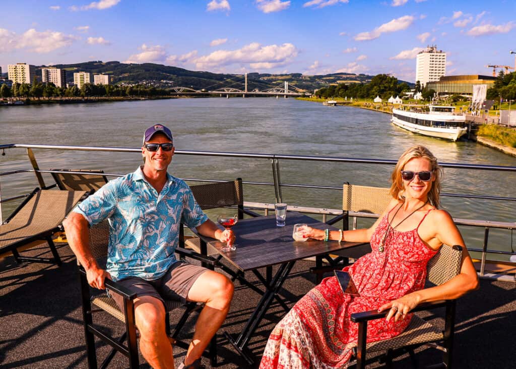 Man and lady having drinks on the deck of a cruise ship with a river behind them