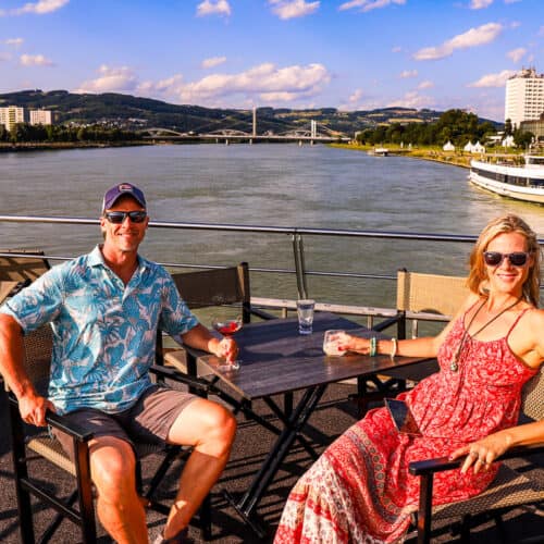 Man and lady having drinks on the deck of a cruise ship with a river behind them