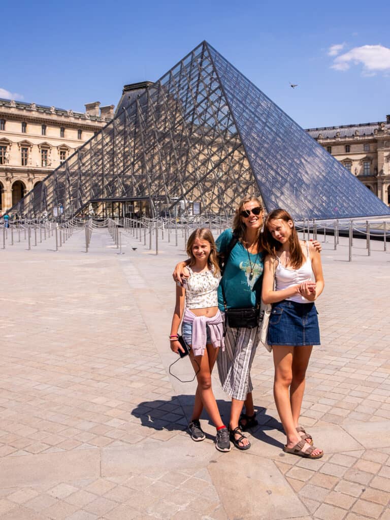 Mom and two daughters standing in front of a glass pyramid at the Louvre museum in Paris