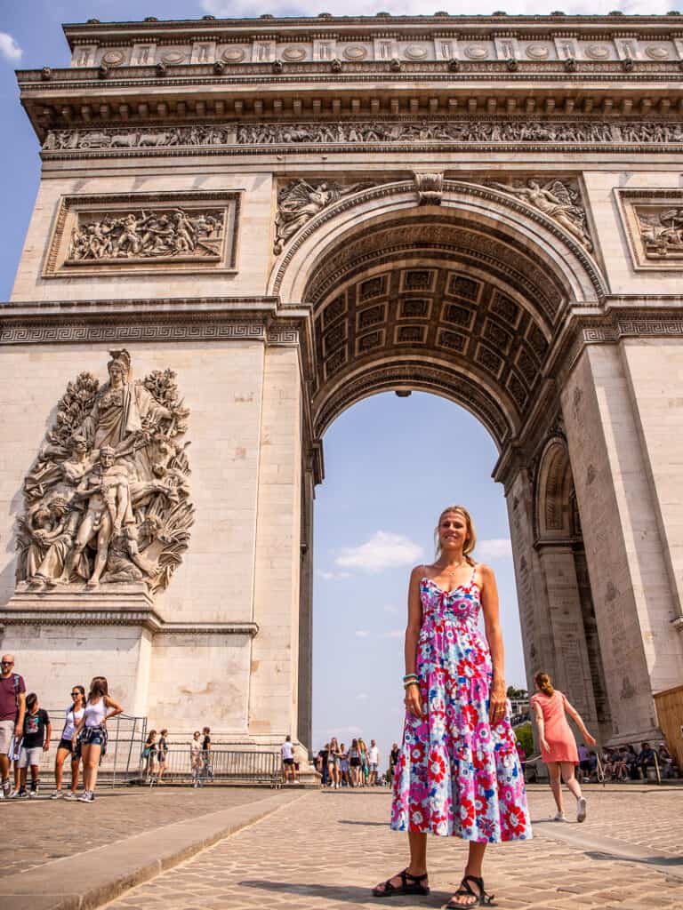 Lady standing under a monument in Paris called Arc De Triomphe