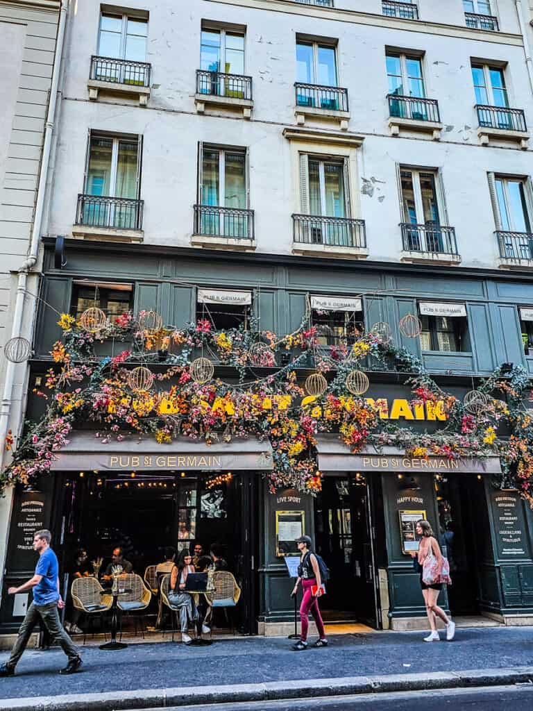 People walking past a pub that's decorated with flowers in Paris