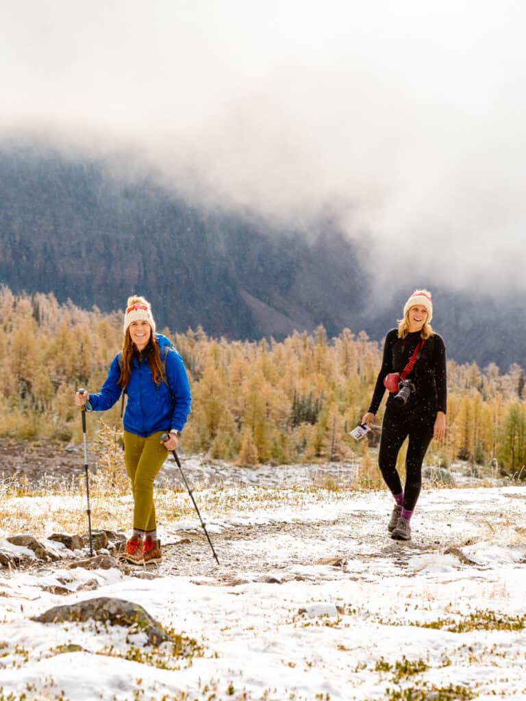 two women hiking on a snow covered trail with larch tree fall foliage