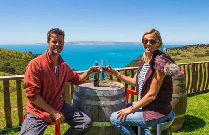 man and woman sitting at wine barrel cheersing with ocean view behind them