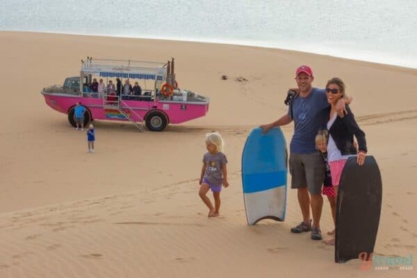 family posing with sandboards on dunes in front of pink lark truck