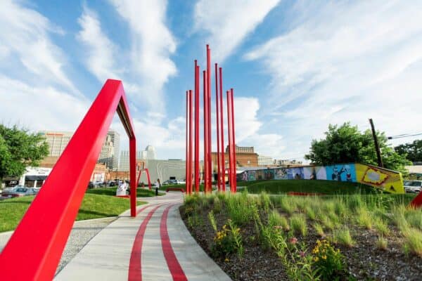 Landscape shot of ARTivity on the Green in Winston-Salem during daytime. Blue skies and greenery with flowers in bloom. Red sculptures in foreground with city in background. Spring or summer season.